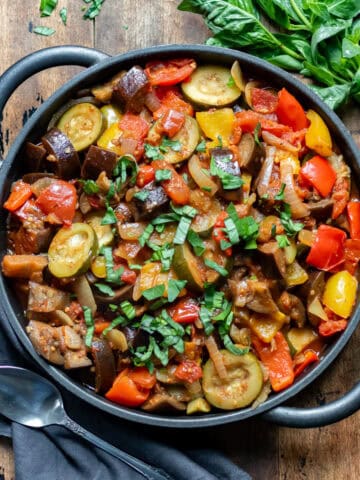 Wooden table with a cast iron serving dish of slow cooker ratatouille, next to a bunch of basil and a napkin.