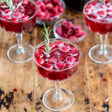 A table with champagne glasses of mulled wine spritzer with frozen cranberries and raspberries and a sprig of rosemary.