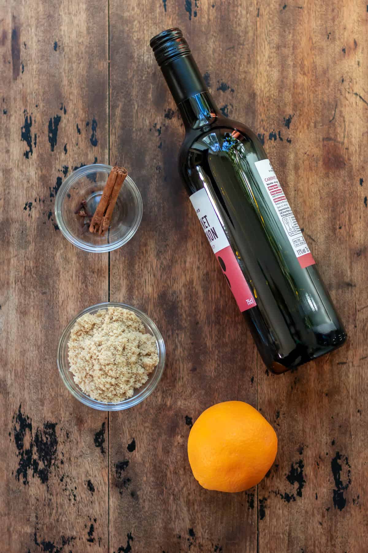 Red wine, brown sugar, an orange, cinnamon stick and whole cloves on a table.