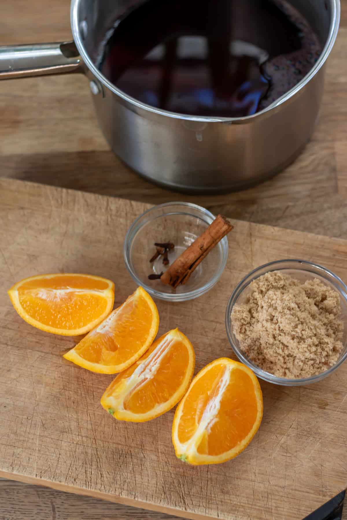 A saucepan of red wine, next to cut oranges, and a dish of sugar, cinnamon and cloves.
