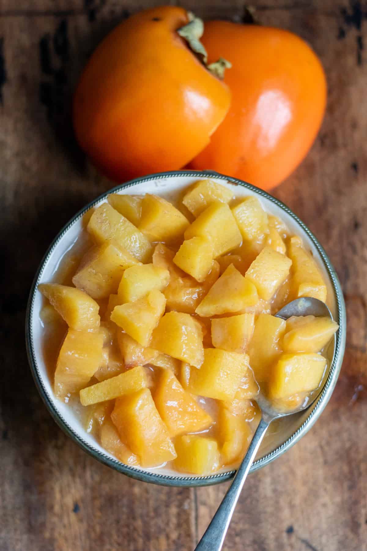 Looking down at a dish of persimmon compote on a table next to two persimmons.