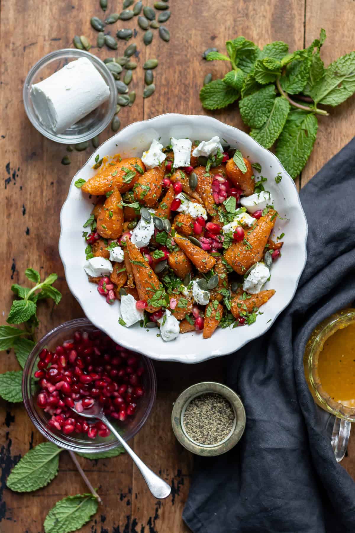Serving dish of roasted carrot salad, next to mint, goat's cheese and pomegranate seeds on the table.
