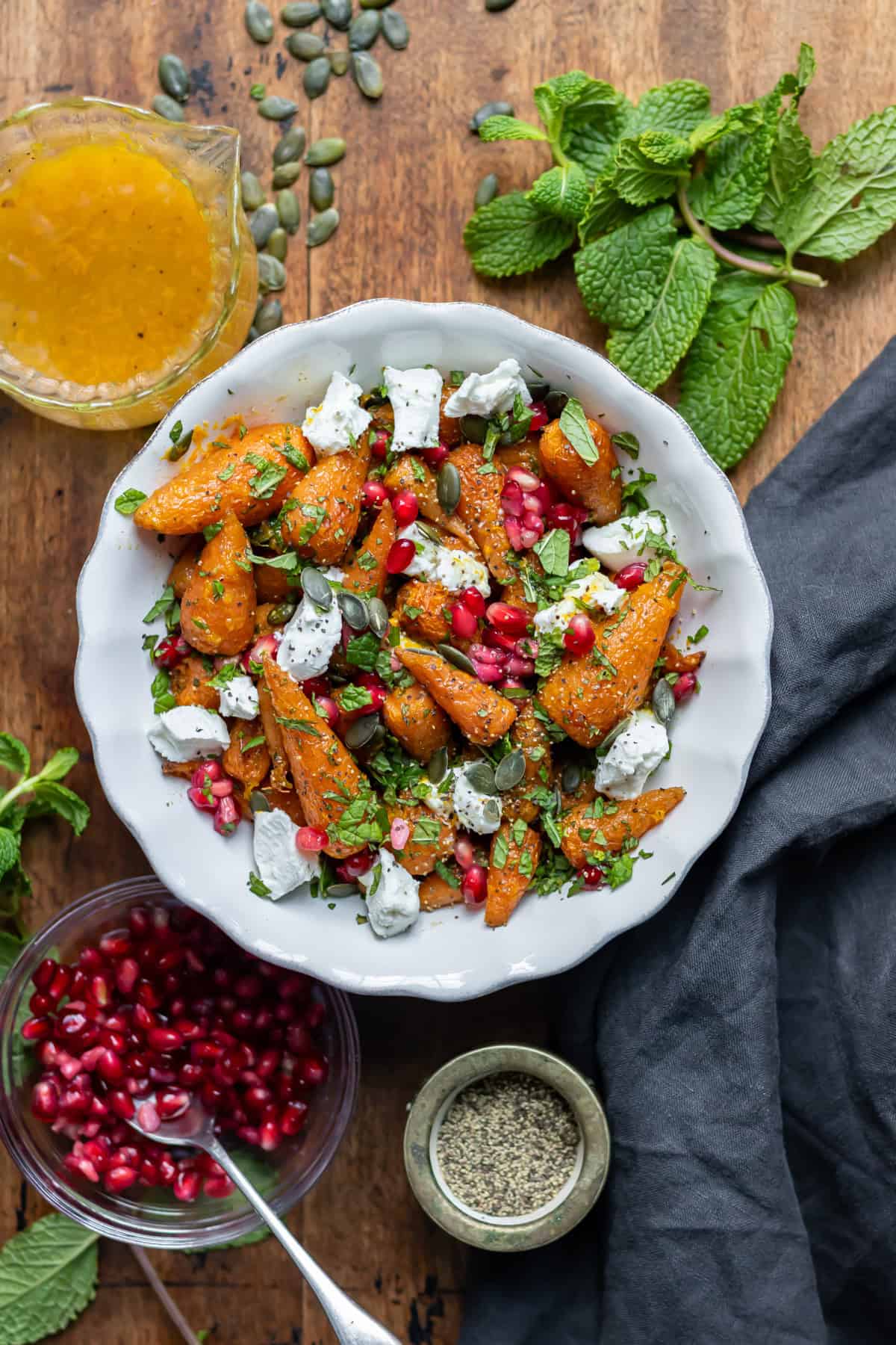 Serving dish of roasted carrot salad, next to mint, goat's cheese and pomegranate seeds on the table.