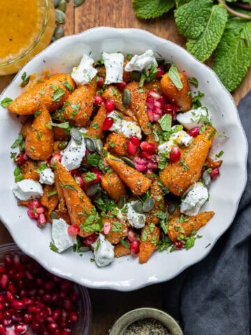 Serving dish of roasted carrot salad, next to mint, goat's cheese and pomegranate seeds on the table.