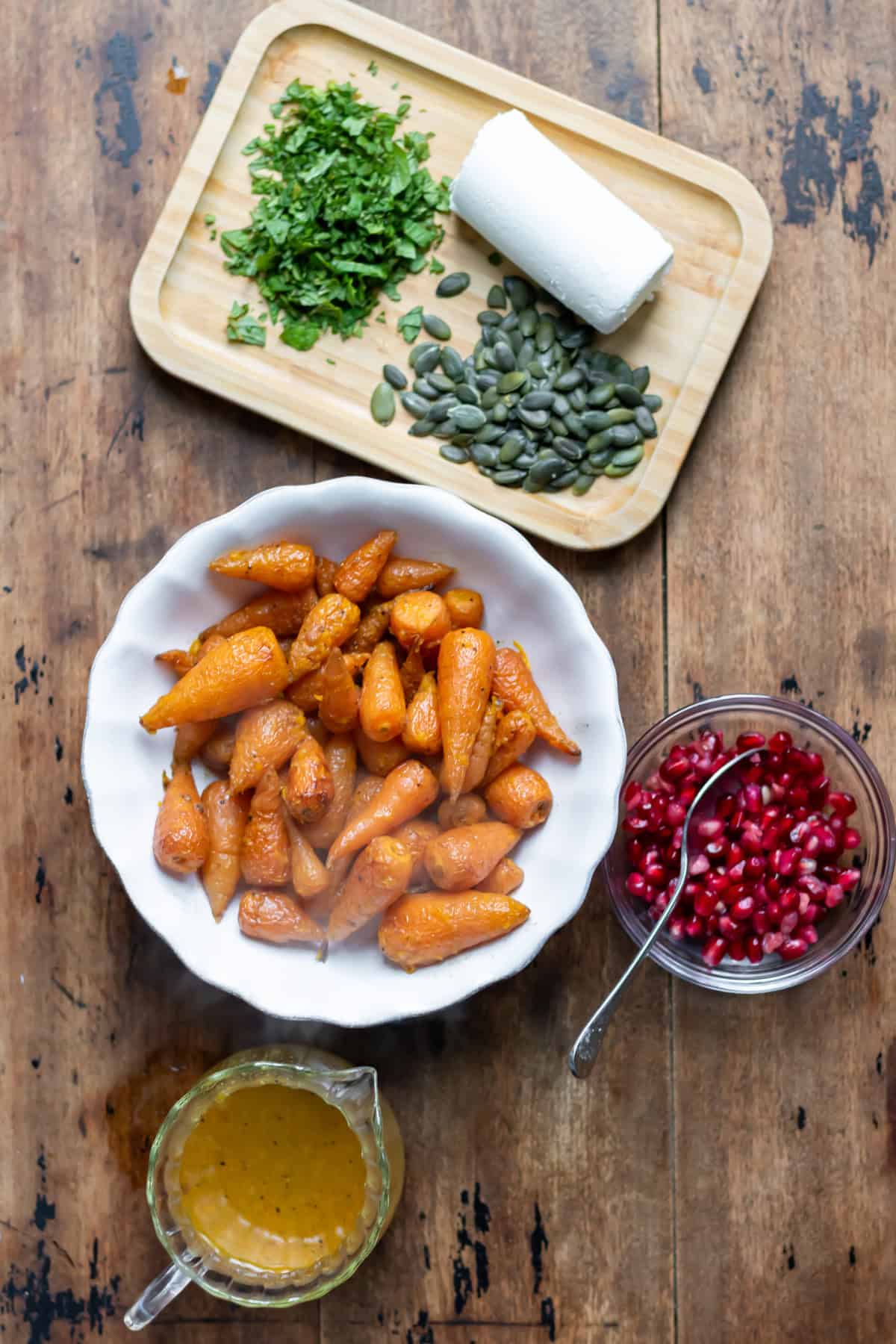 Table with the roasted carrots in orange dressing, next to pomegranate seeds, pumpkin seeds, chopped mint and goat's cheese.