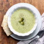 Looking down at a wooden table with a bowl of blended broccoli soup with crackers next to it.