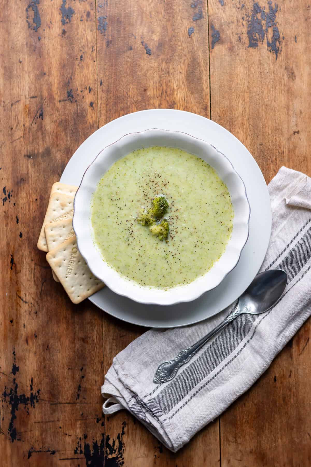 Looking down at a wooden table with a bowl of blended broccoli soup with crackers next to it.