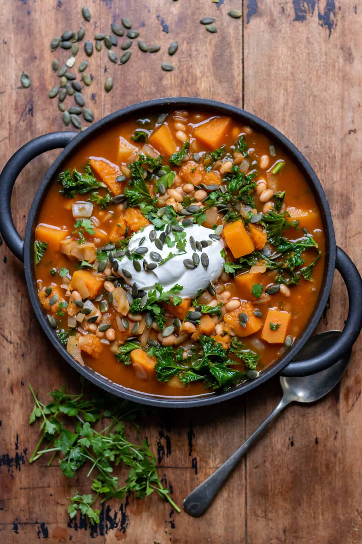Wooden table with a serving dish of kale butternut squash and bean soup, topped with yogurt, herbs and pumpkin seeds.