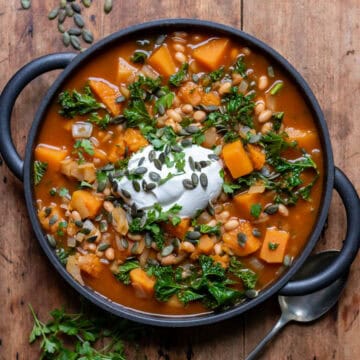 Wooden table with a serving dish of kale butternut squash and bean soup, topped with yogurt, herbs and pumpkin seeds.