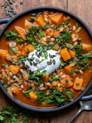 Wooden table with a serving dish of kale butternut squash and bean soup, topped with yogurt, herbs and pumpkin seeds.