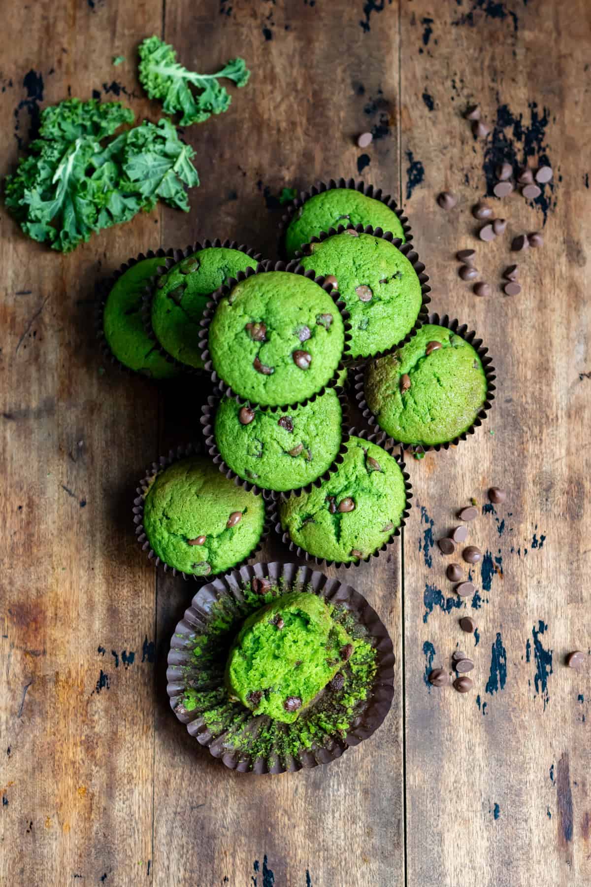 Looking down at a wooden table with a pile of kale muffins, one with a bite out, next to some kale and chocolate chips.