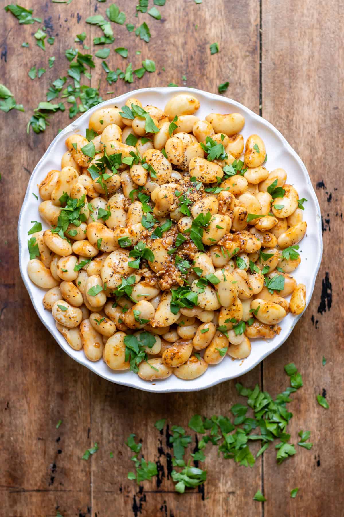 A wooden table with a plate of smoky garlic butter beans topped with fresh parsley.