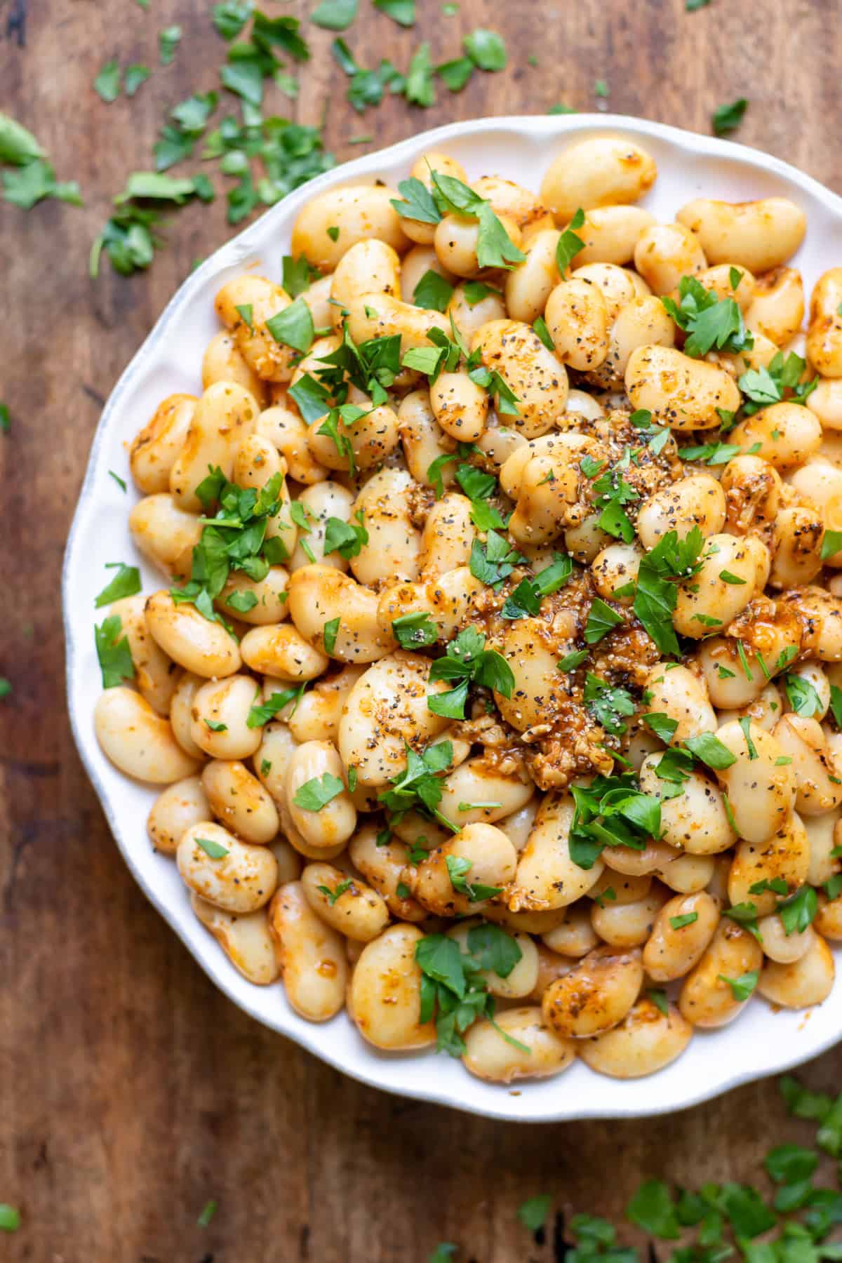 A wooden table with a plate of smoky garlic butter beans topped with fresh parsley.
