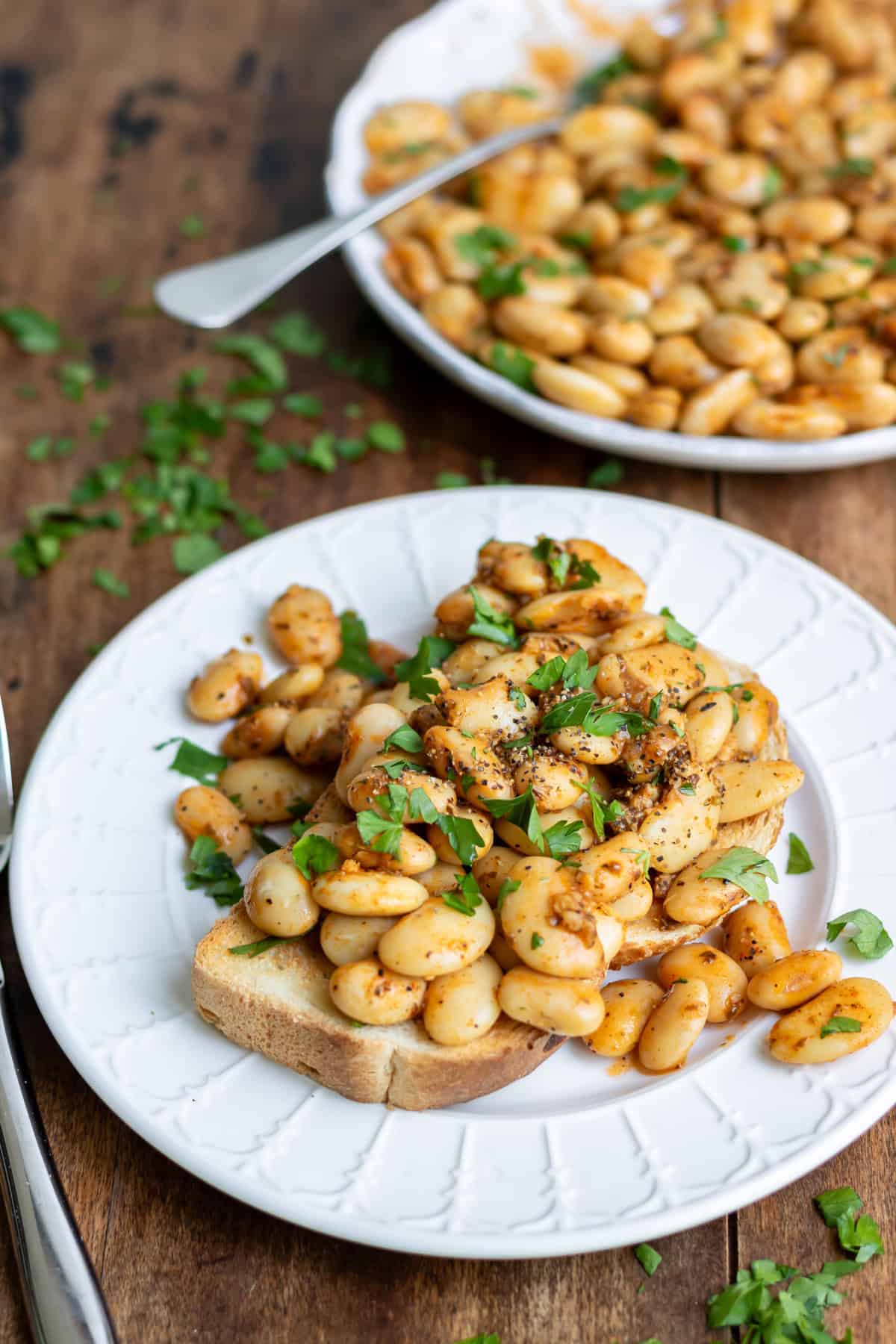 A plate with garlic butter beans on toasted sourdough, next to a serving dish of more garlic butterbeans.