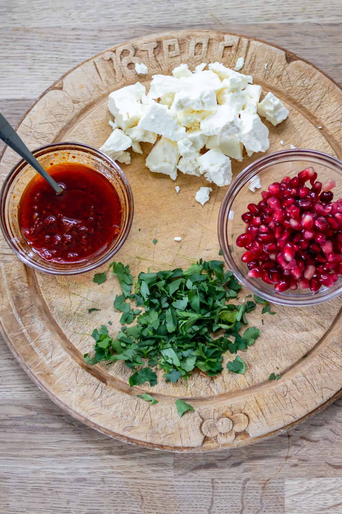A wooden board with a pile of crumbled feta, dish of pomegranate seeds, chopped parsley and the harissa-infused oil.