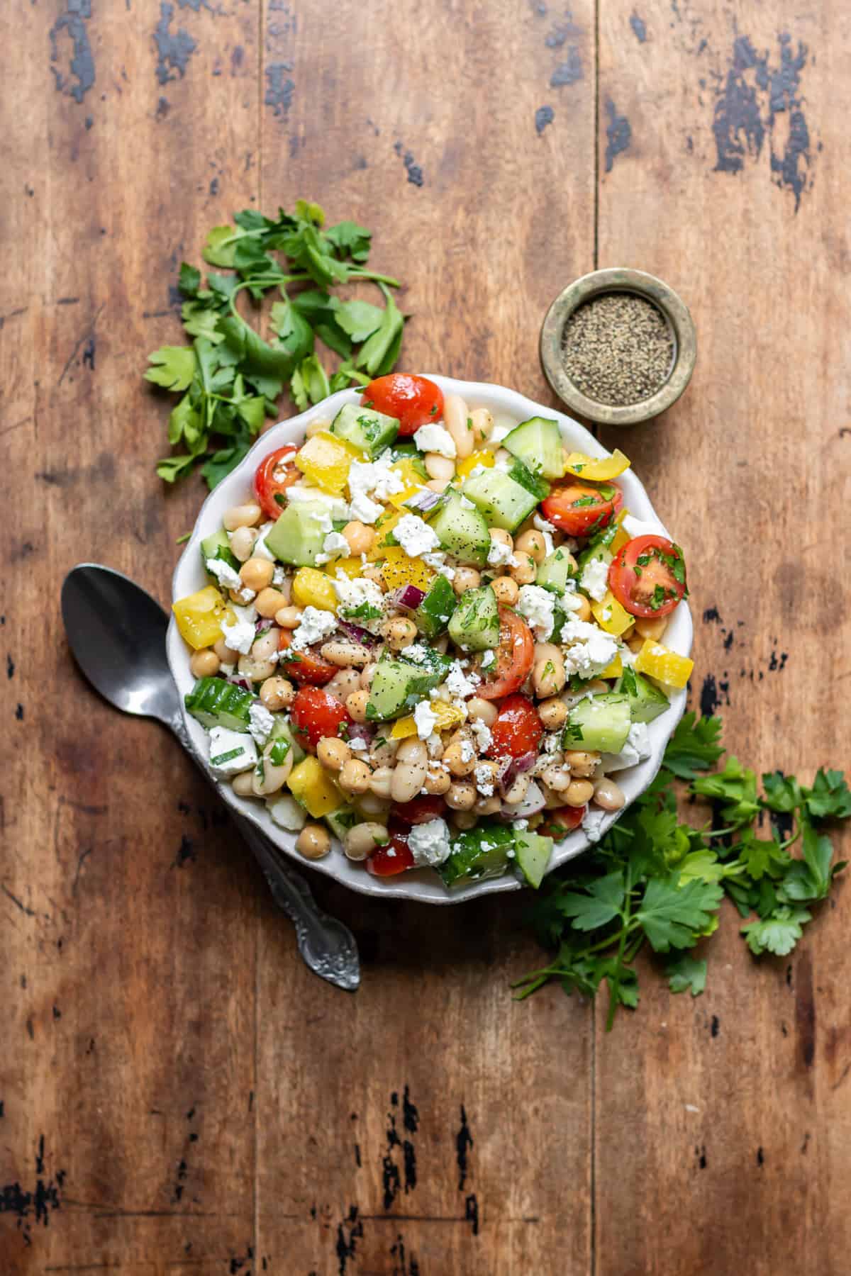 A wooden table with a serving dish of Mediterranean Bean Salad with some sprigs of parsley next to it.