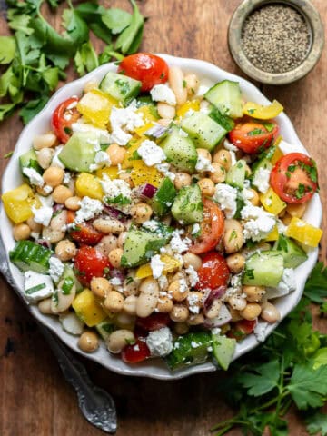 A wooden table with a serving dish of Mediterranean Bean Salad with some sprigs of parsley next to it.