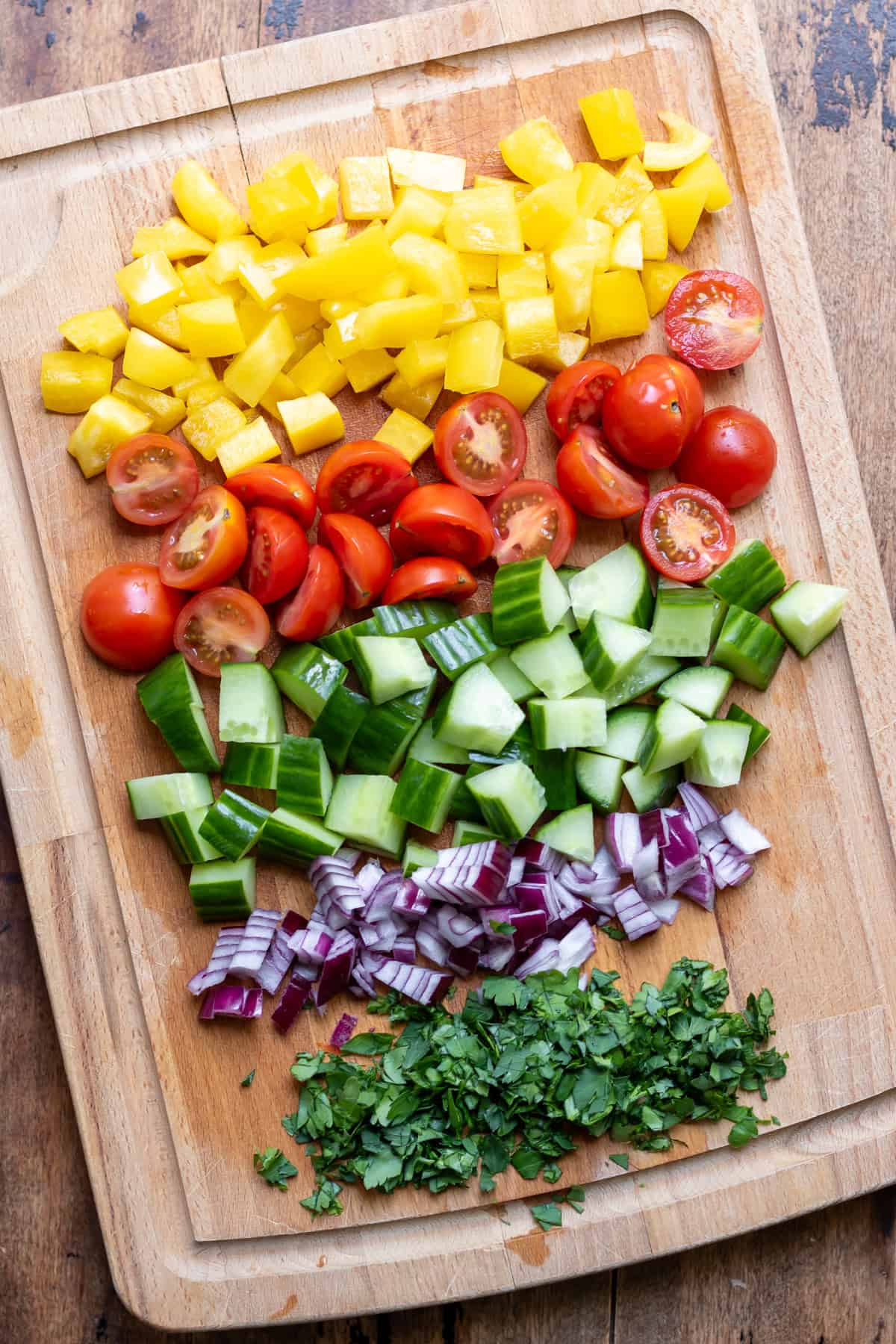 A wooden cutting board with diced yellow bell pepper, halved cherry tomatoes, diced cucumber, diced red onion and chopped parsley.