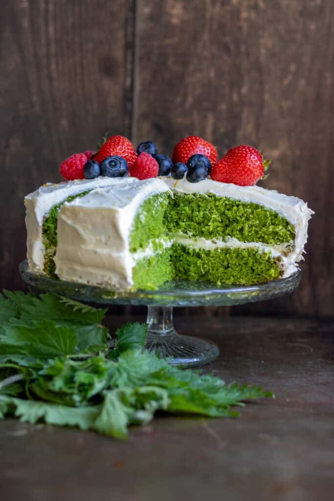 A stinging nettle layer cake with a slice removed, on a glass cake stand.