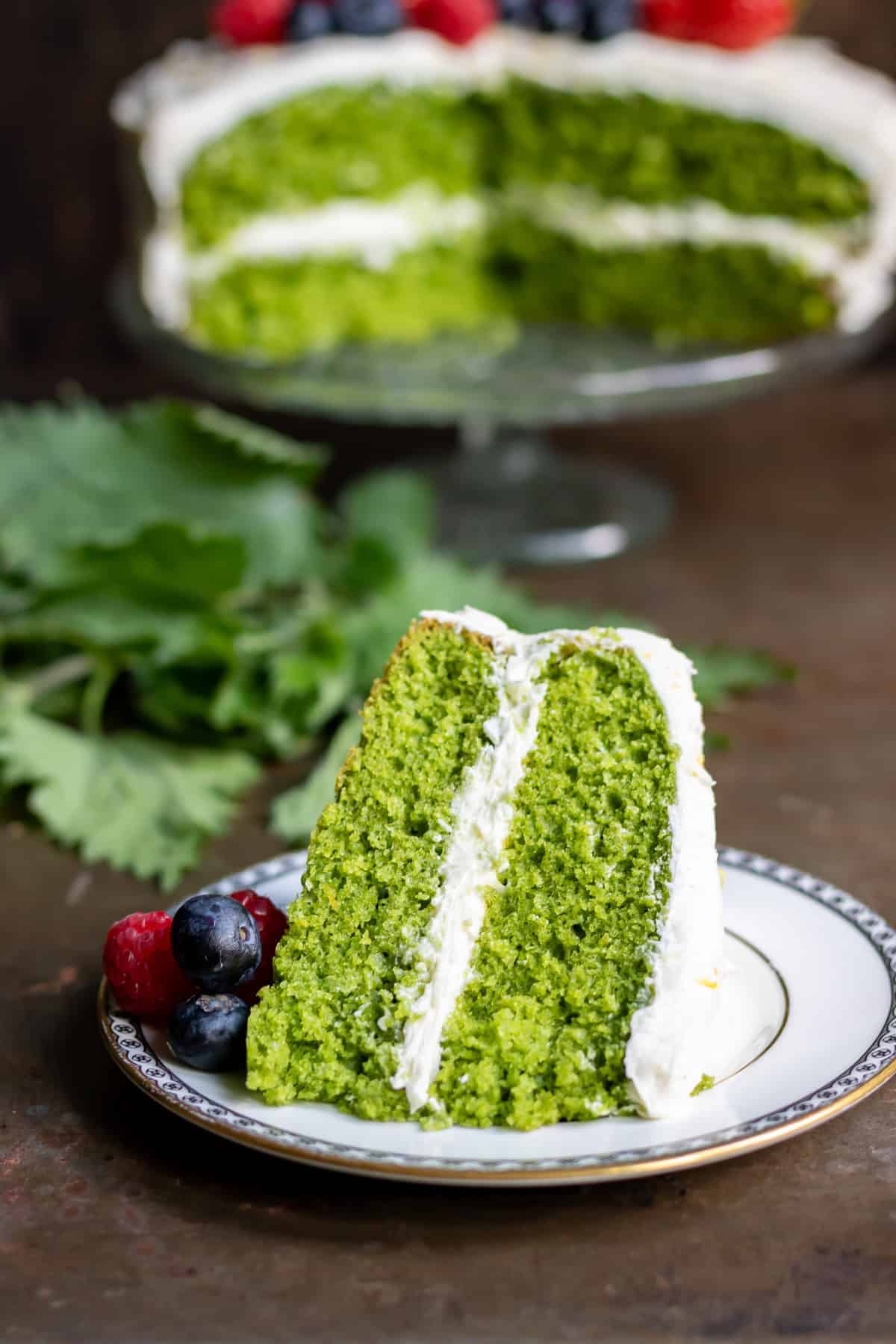 A slice of stinging nettle cake on a plate, in front of the rest of the cake on a glass cake stand.