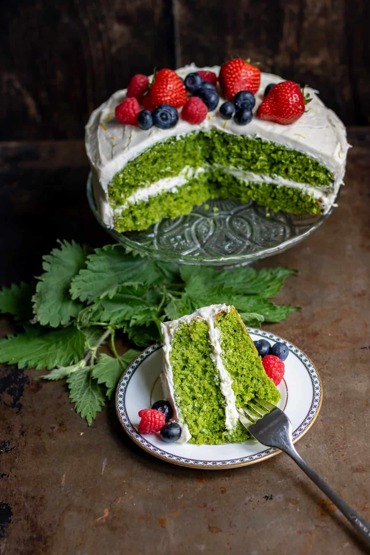 Looking down at a table with a stinging nettle cake on a cake stand, with a slice of it on a plate in front.
