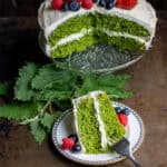 Looking down at a table with a stinging nettle cake on a cake stand, with a slice of it on a plate in front.