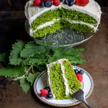 Looking down at a table with a stinging nettle cake on a cake stand, with a slice of it on a plate in front.