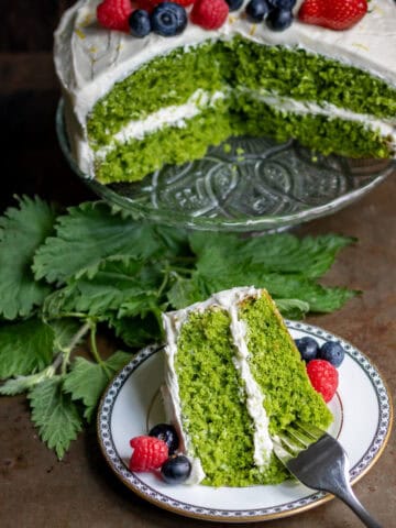 Looking down at a table with a stinging nettle cake on a cake stand, with a slice of it on a plate in front.