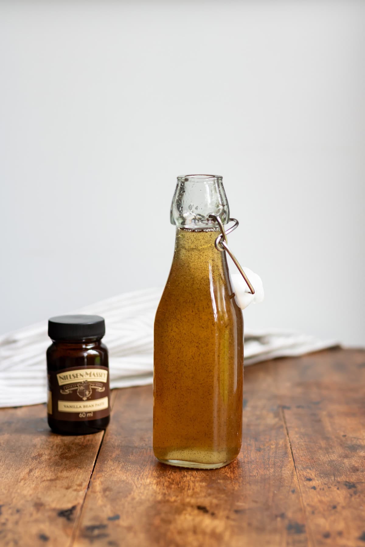 Wooden table with a glass bottle of vanilla simple syrup, jar of vanilla bean paste and a cloth napkin.