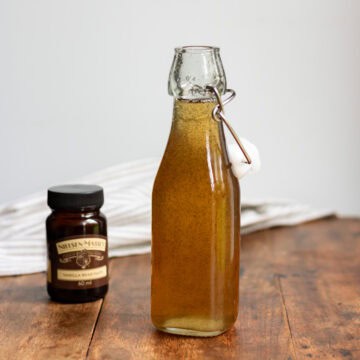 Wooden table with a glass bottle of vanilla simple syrup, jar of vanilla bean paste and a cloth napkin.