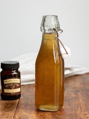 Wooden table with a glass bottle of vanilla simple syrup, jar of vanilla bean paste and a cloth napkin.