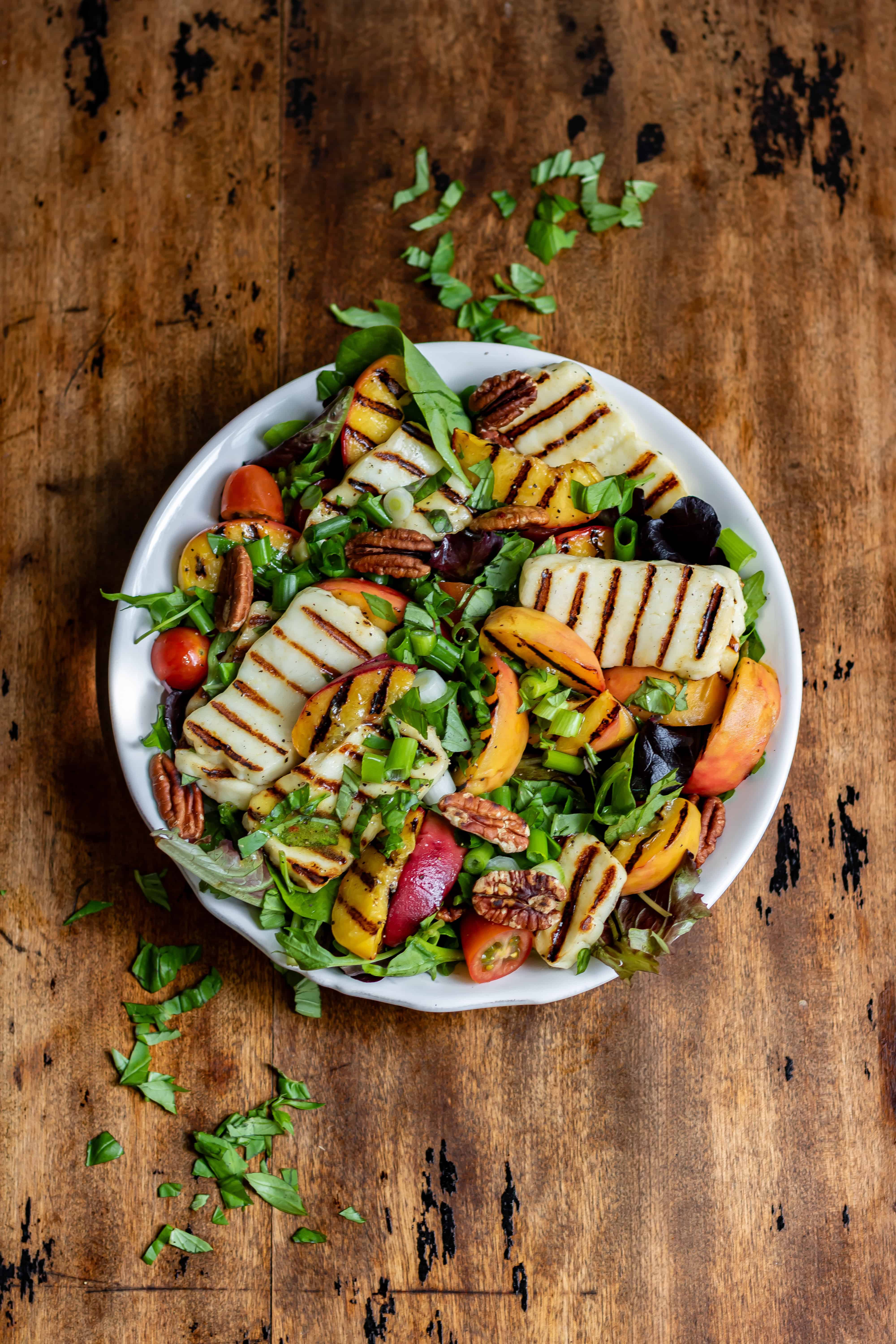 Looking down at a wooden table with a bowl of grilled peach halloumi salad with hot honey vinaigrette.