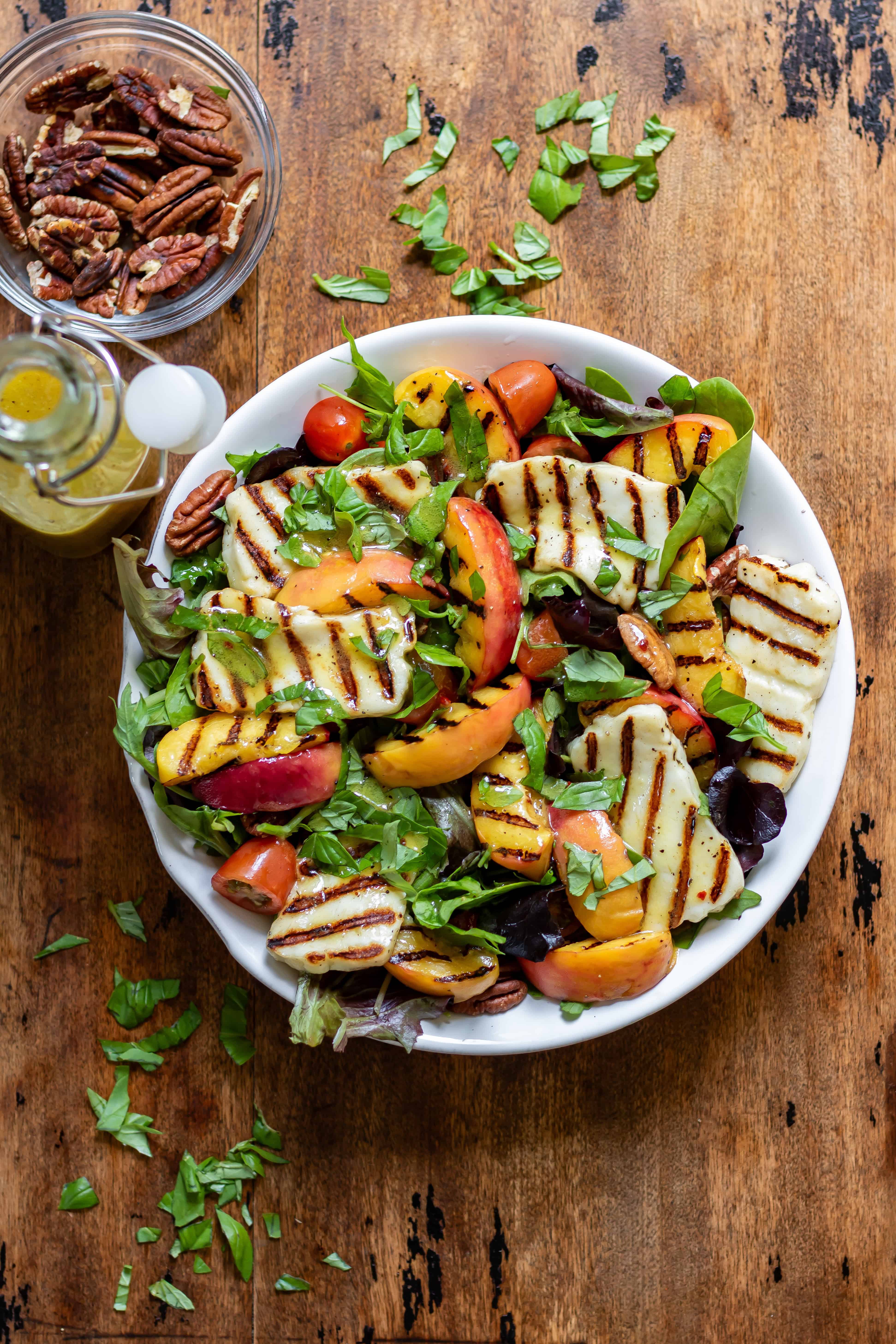 Wooden table with a serving dish of grilled halloumi peach salad, with a dish of toasted pecans next to it, and a bottle of hot honey vinaigrette.