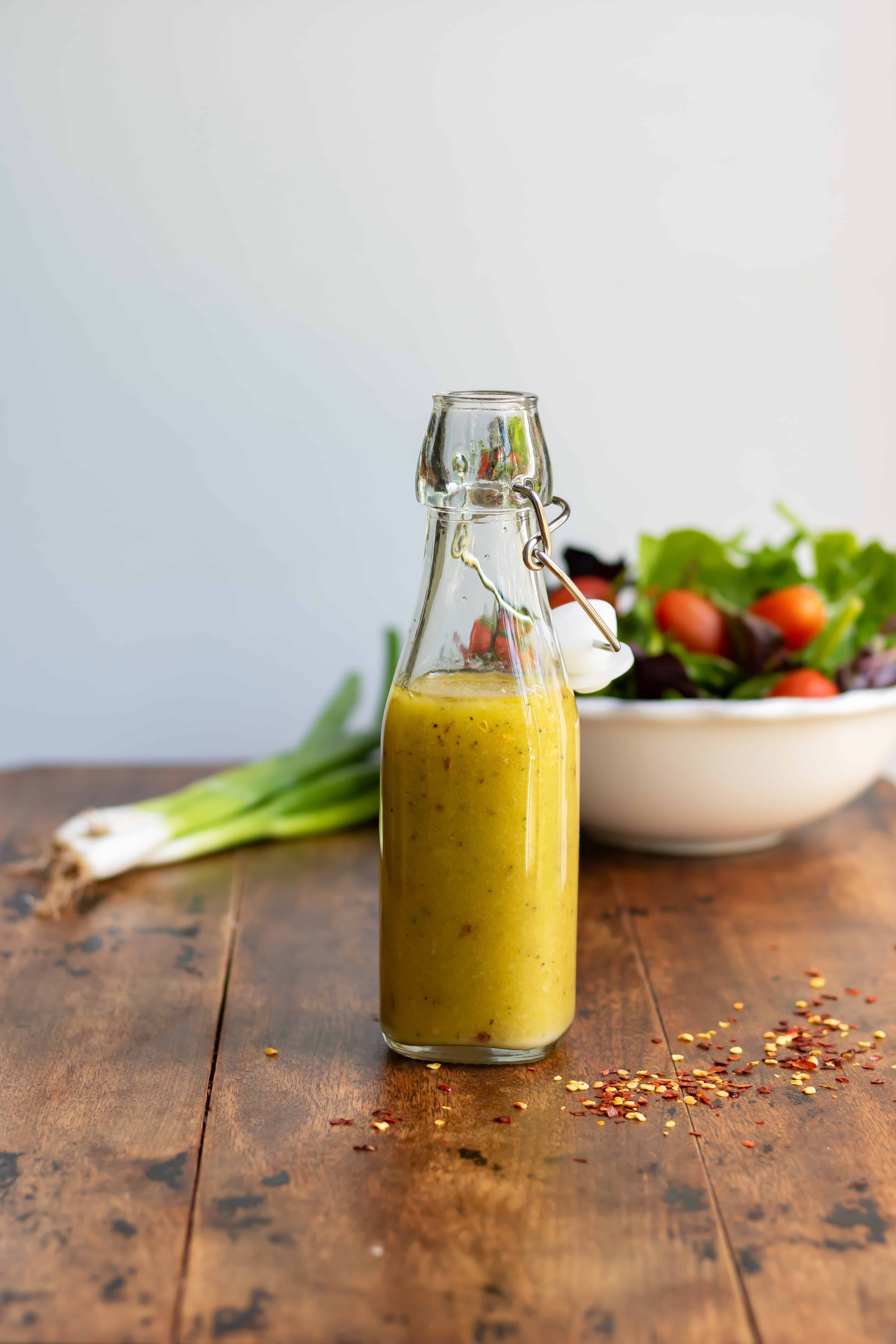 Wooden table with a dish of salad and some spring onions, with a glass bottle of hot honey vinaigrette.