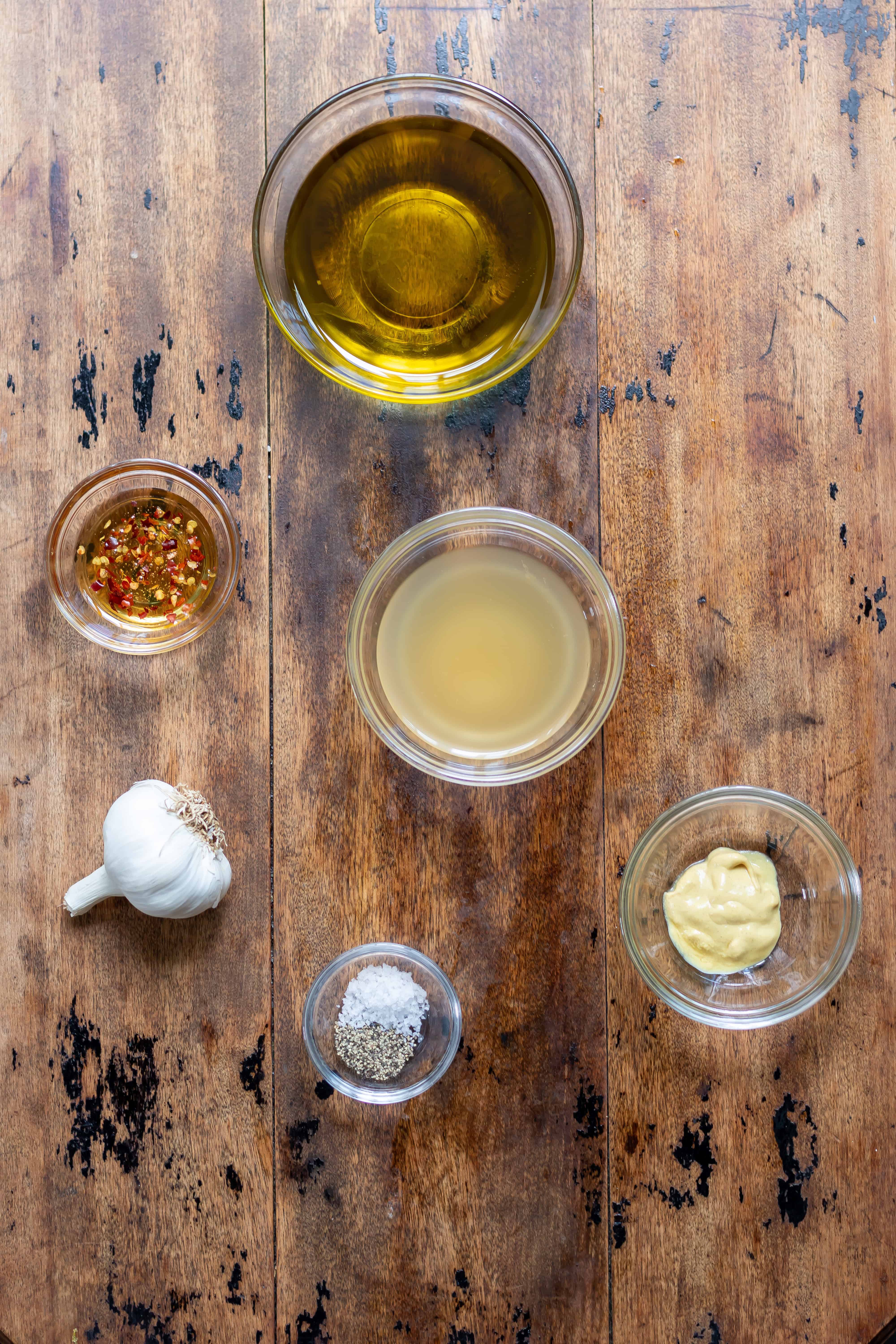 Small glass bowls of extra virgin olive oil, apple cider vinegar, dijon mustard, hot honey, salt and pepper, and a bulb of garlic.