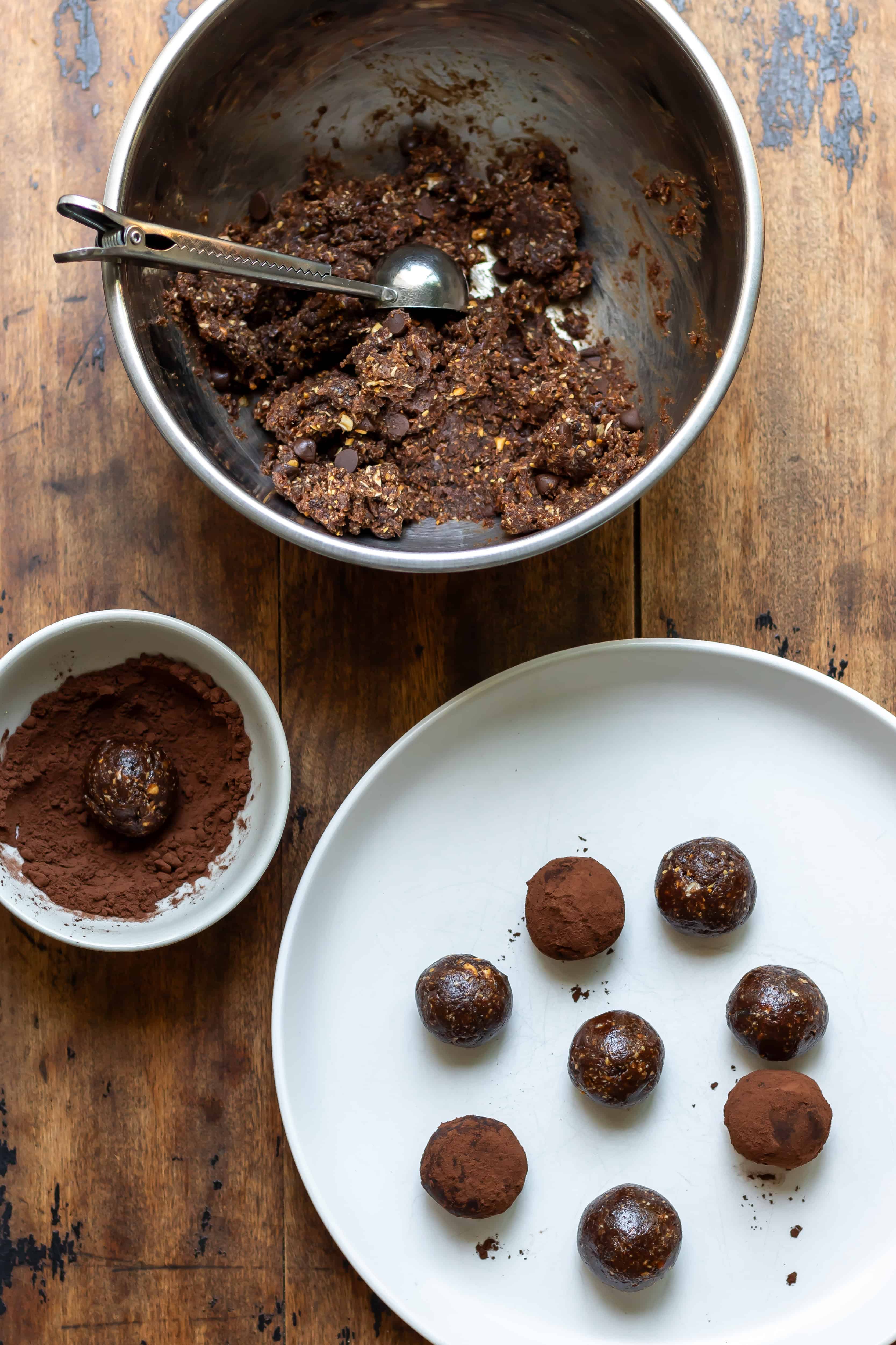 Bowl of protein ball mixture, one being rolled in cocoa powder, and some finished ones on a plate.