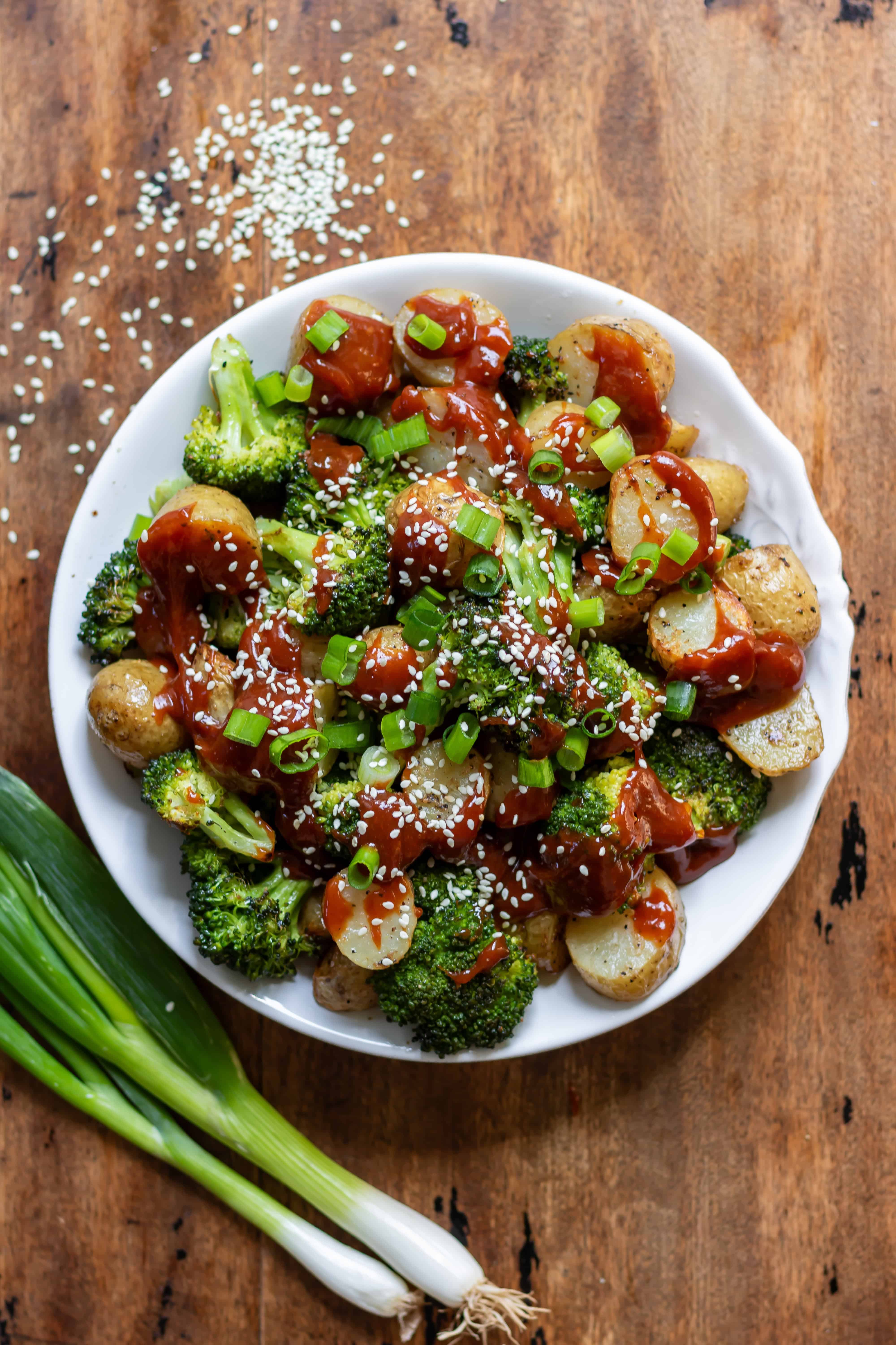 A wooden table with a serving dish of Koran Gochujang Roasted Potatoes and Broccoli.