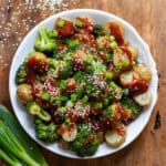 Looking down at a dish of roasted broccoli and potatoes with Korean gochujang sauce, spring onions and sesame seeds.
