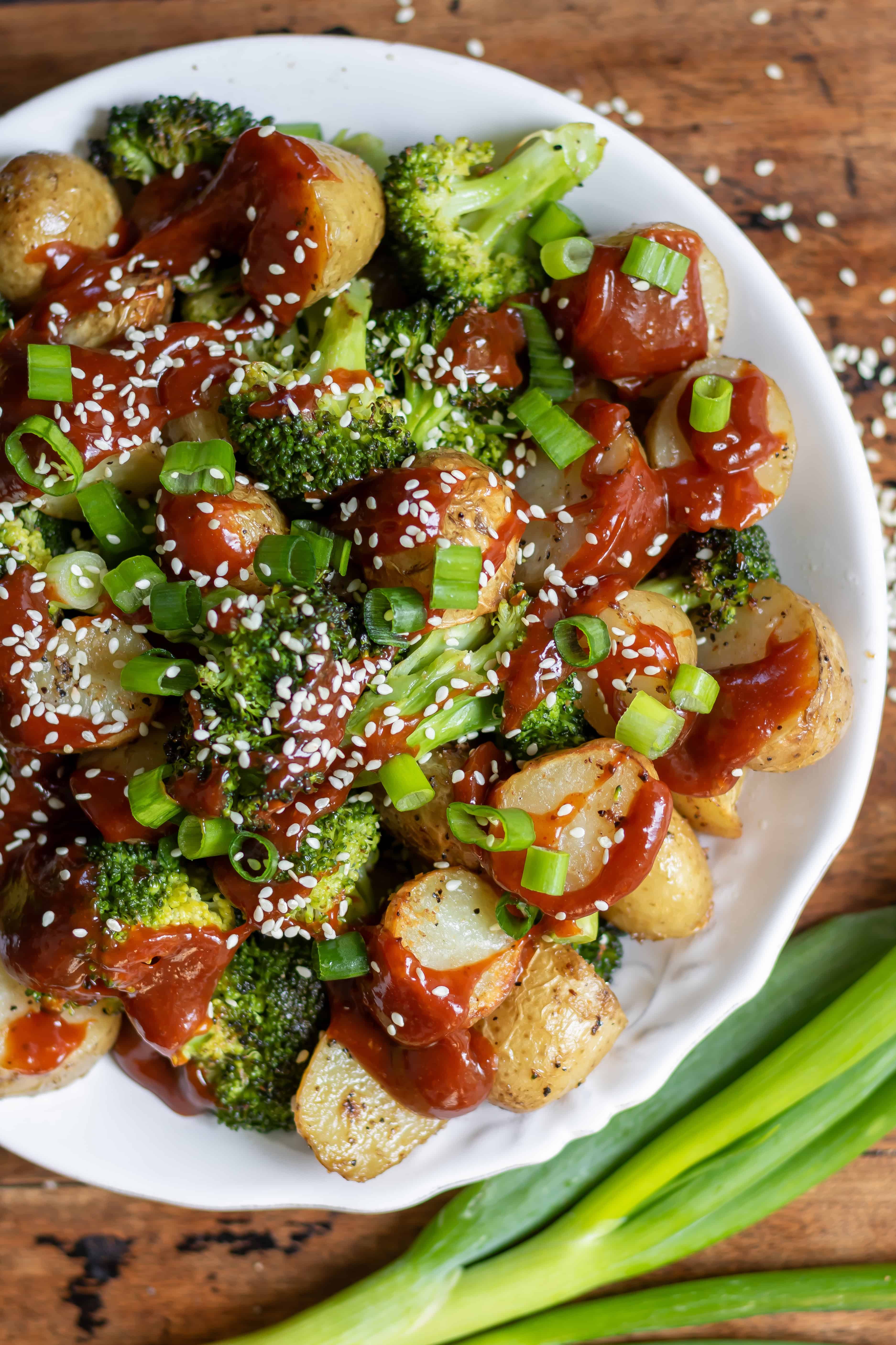 Looking down at a dish of roasted broccoli and potatoes with Korean gochujang sauce, spring onions and sesame seeds.