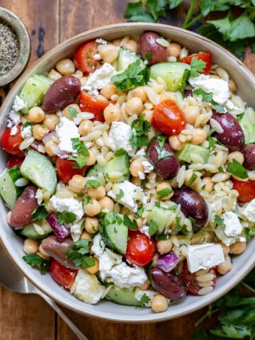 Wooden table with a serving dish of Mediterranean Orzo Salad with sprigs of parsley next to it.