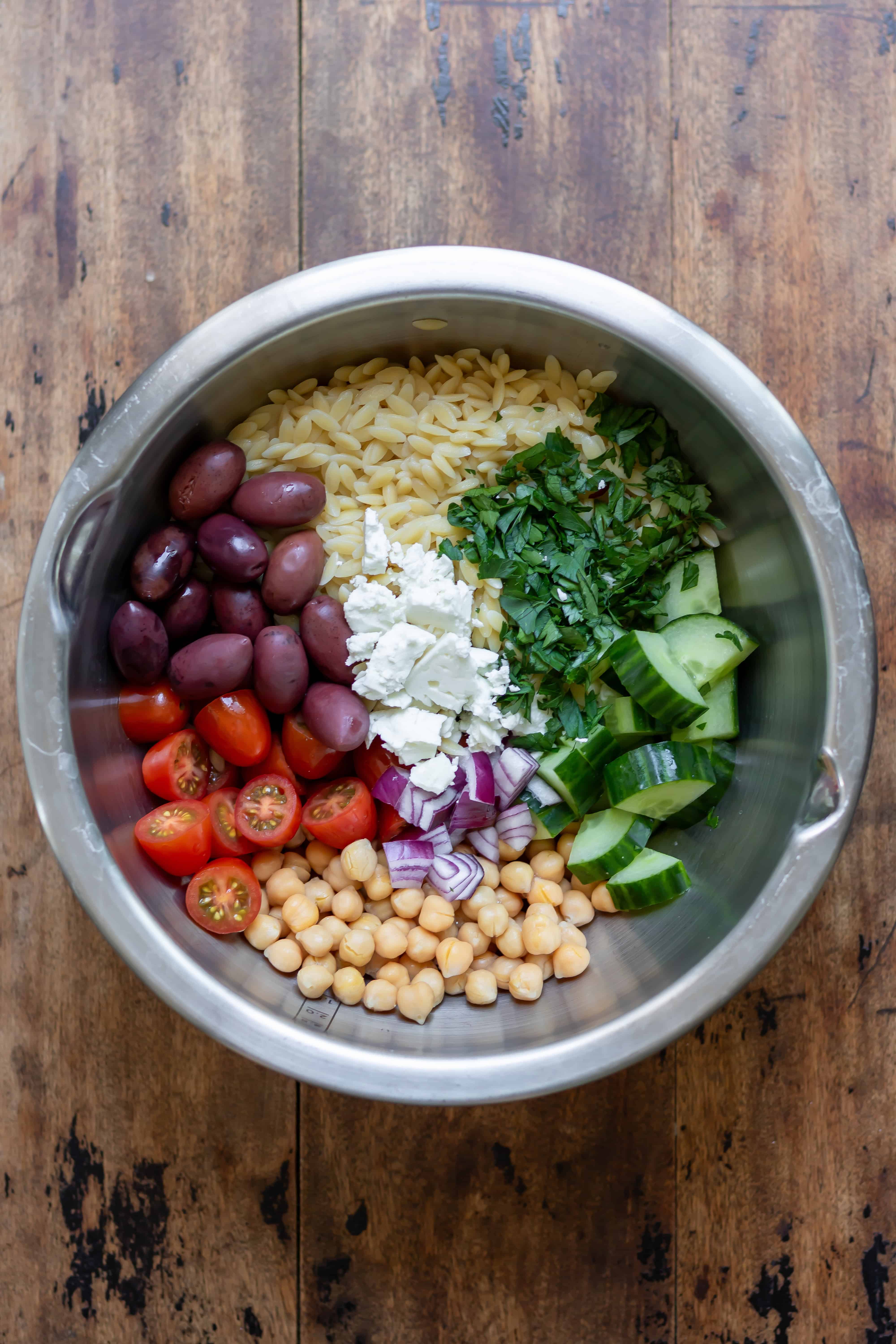 Metal mixing bowl with cooked orzo, chickpeas, veggies, olives and feta.