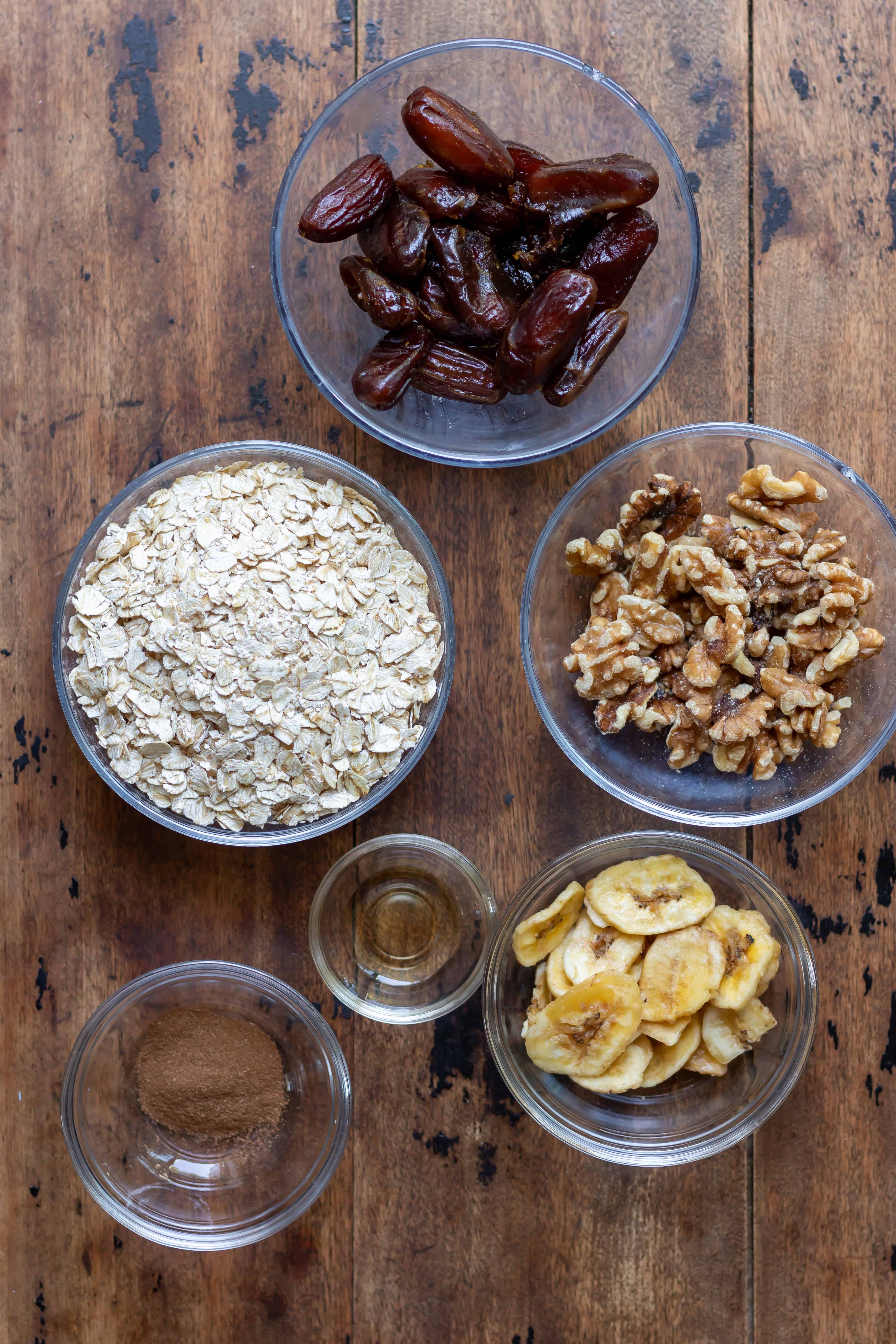 Dates, oats, walnuts, cinnamon, vanilla and dried banana chips in dishes on a table.