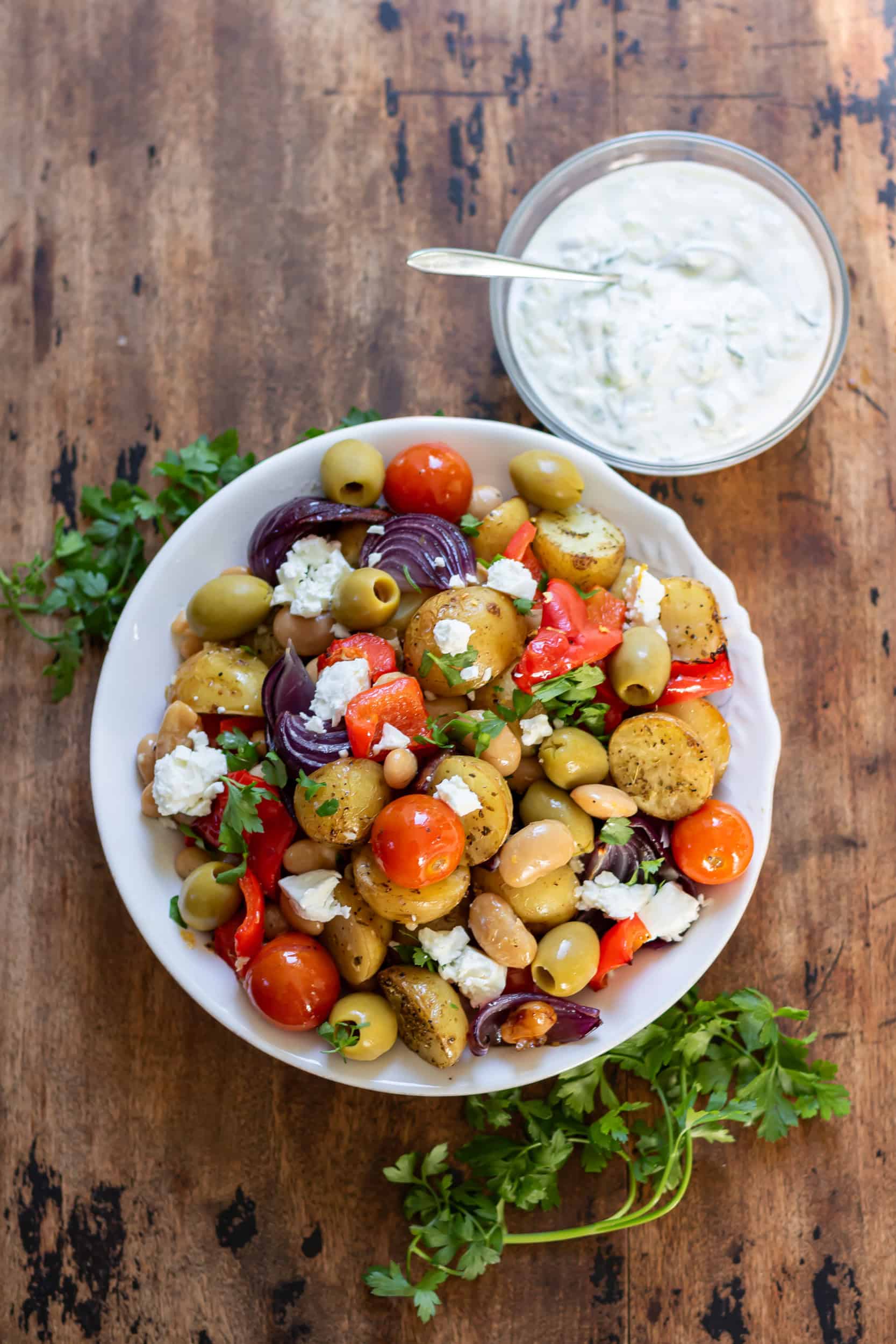 Looking down at a dish of Greek Butter Bean and Potato Tray Bake next to a bowl of tzatziki sauce.