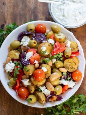 Looking down at a dish of Greek Butter Bean and Potato Tray Bake next to a bowl of tzatziki sauce.
