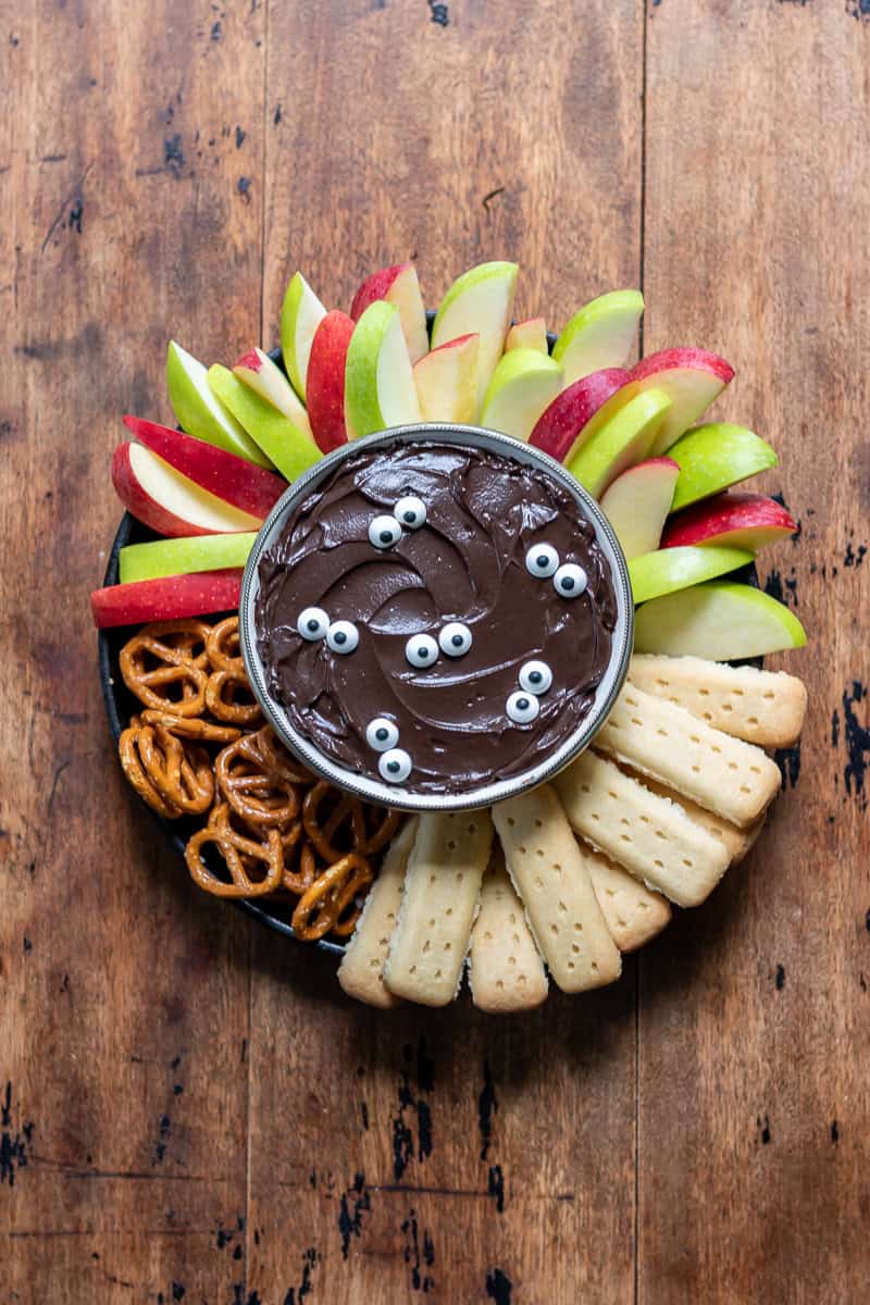 A wooden table with a bowl of halloween black cocoa cheesecake dip topped with candy eyes and surrounded by sliced apples, pretzels and shortbread.