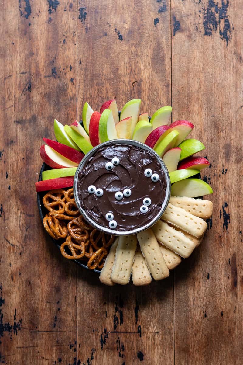 A wooden table with a bowl of halloween black cocoa cheesecake dip topped with candy eyes and surrounded by sliced apples, pretzels and shortbread.