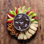 A wooden table with a bowl of halloween black cocoa cheesecake dip topped with candy eyes and surrounded by sliced apples, pretzels and shortbread.
