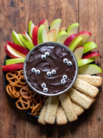 A wooden table with a bowl of halloween black cocoa cheesecake dip topped with candy eyes and surrounded by sliced apples, pretzels and shortbread.
